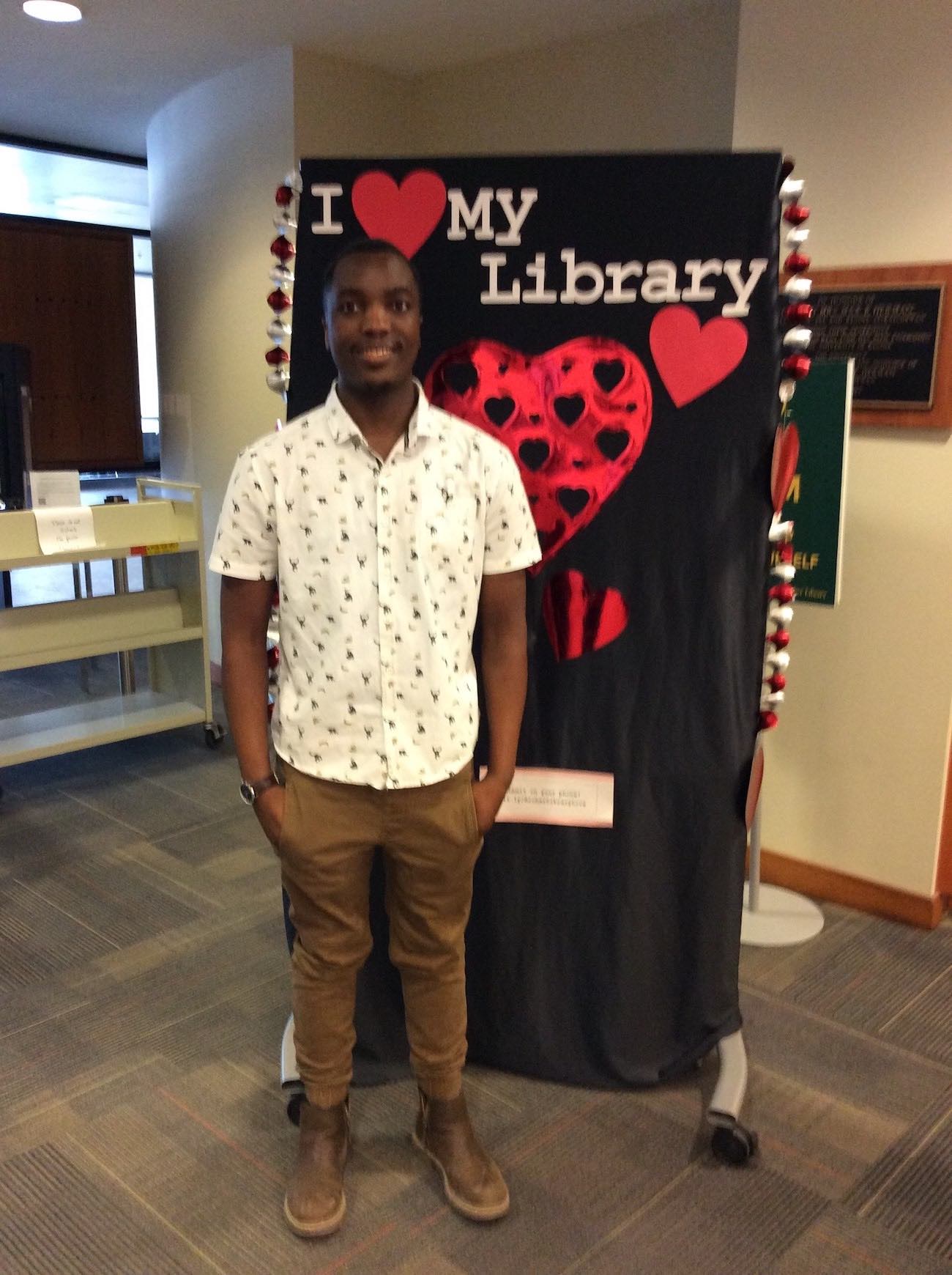 A smiling student with his hands in his pockets stands in front of the I love my library backdrop